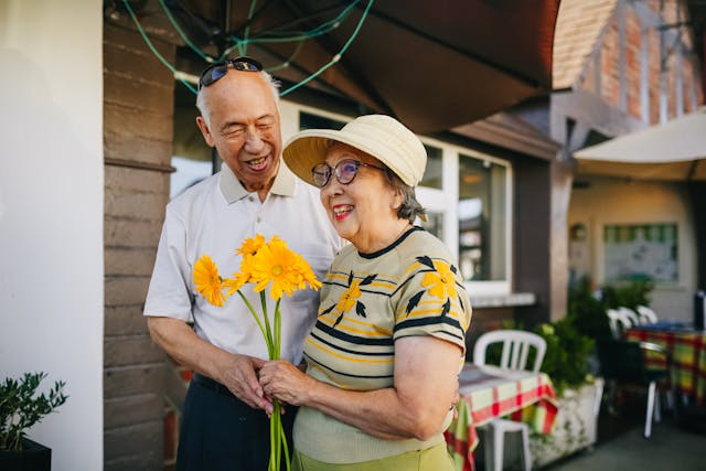 Older man and woman smiling and standing outside, the woman is holding yellow flowers